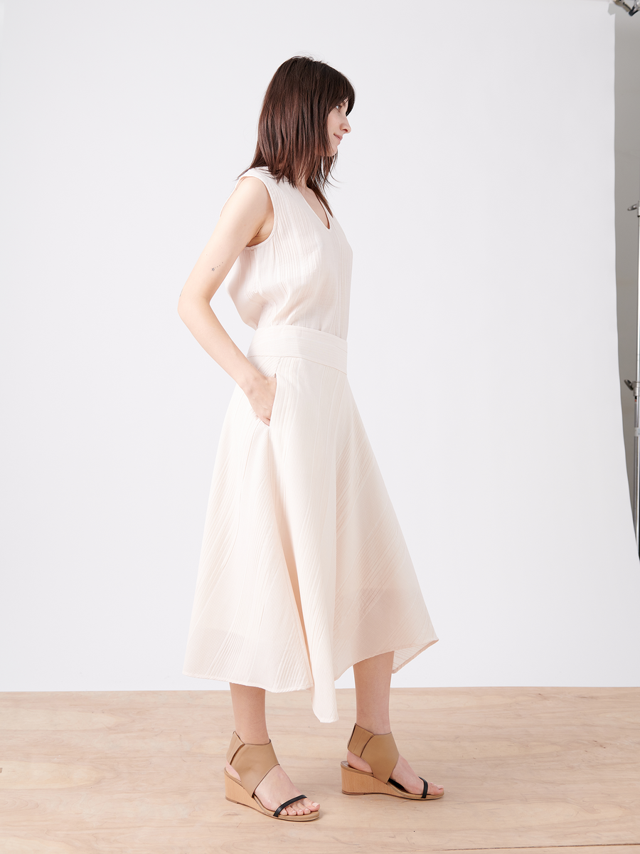 A woman with shoulder-length hair stands sideways in the Aki Wave Skirt by Zero + Maria Cornejo, a sleeveless off-white dress with an asymmetrical skirt. She pairs it with tan sandals and rests her left hand in the pocket against a white background, highlighting her elegant ensemble.