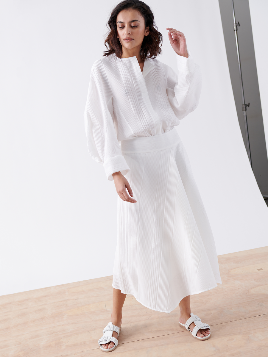 In a minimalist studio, a woman stands on a wooden floor against a white backdrop, wearing Zero + Maria Cornejo's Aki Wave Skirt - Final Sale, paired with a loose-fitting white blouse and white sandals, looking slightly downward.