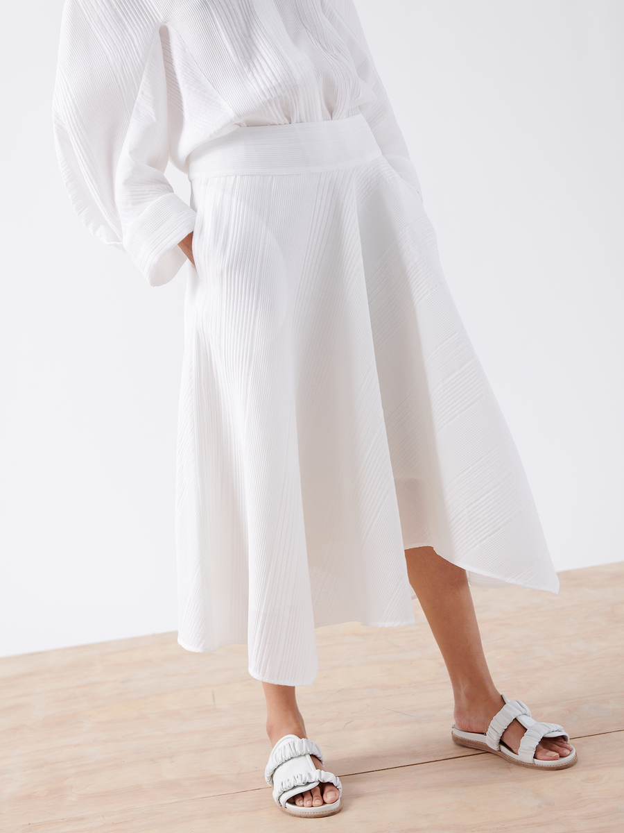 A person is wearing the Aki Wave Skirt - Final Sale by Zero + Maria Cornejo, a white pleated midi skirt with a banded waist, paired with a matching white top and textured slide sandals. The outfit, set against a minimal light background, showcases clean and elegant style.