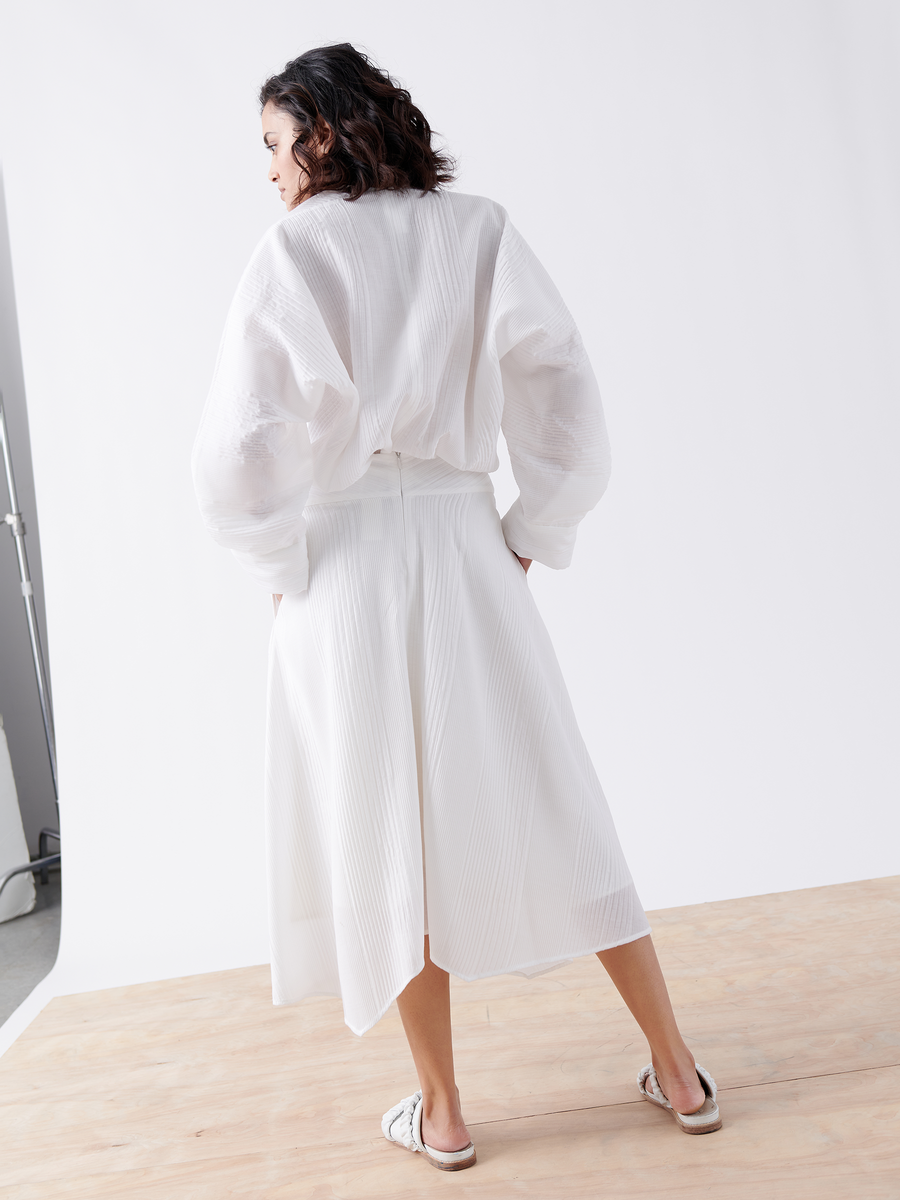 A woman stands on a wooden floor against a white backdrop, wearing a loose-fitting white blouse and the Aki Wave Skirt by Zero + Maria Cornejo. Her dark, wavy hair cascades down as she faces away from the camera, with slip-on sandals completing her effortlessly elegant look.