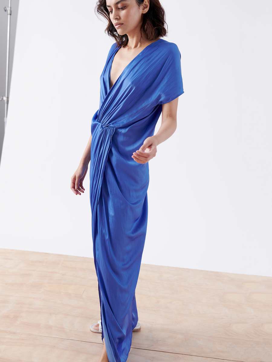 A woman in a Twist Front Dress - Final Sale by Zero + Maria Cornejo, featuring a blue washed georgette and v-neck with twisted detailing, stands on a wooden floor against a white background. Her head tilts slightly as she gracefully extends one hand while looking down.