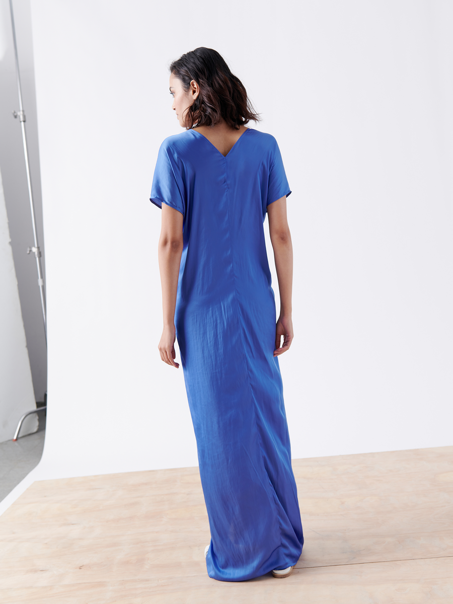 A woman with shoulder-length dark hair stands on a wooden floor against a white backdrop, wearing the Twist Front Dress by Zero + Maria Cornejo. The washed georgette dress is blue silk with a V-shaped back, twisted v-neck detailing, and short sleeves.