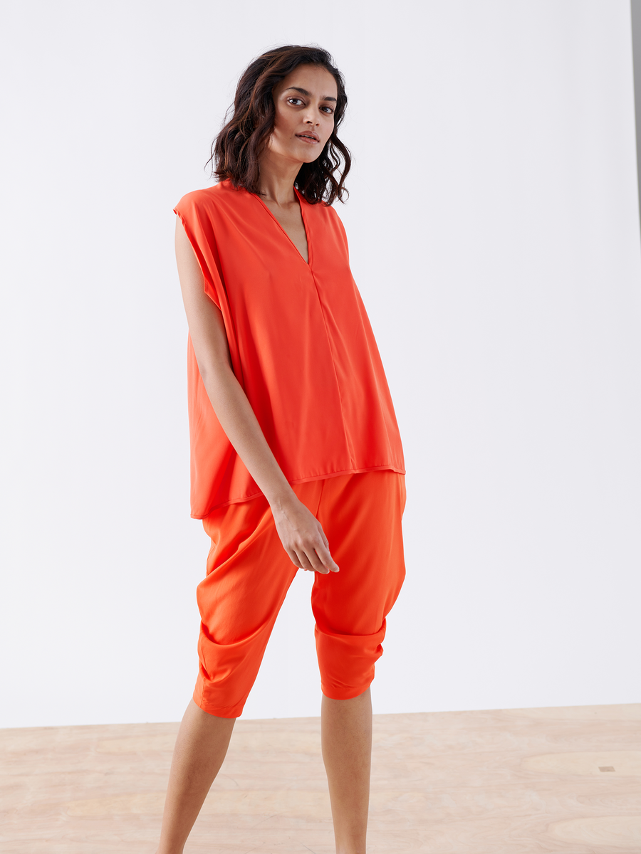 A person with shoulder-length wavy hair wears a loose-fitting, bright orange sleeveless top and the Crop Curve Ori Pant - Final Sale from Zero + Maria Cornejo. Posing confidently, they stand on a wooden floor against a plain white backdrop.