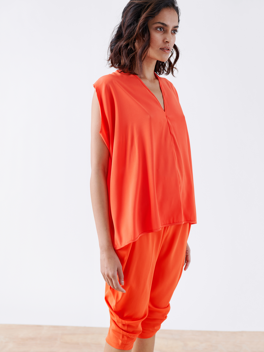 A person in a bright orange sleeveless top and Zero + Maria Cornejo's Crop Curve Ori Pants, made from silk charmeuse with an elastic waist, stands against a plain white background. They have shoulder-length wavy hair, showcasing a style crafted in New York.