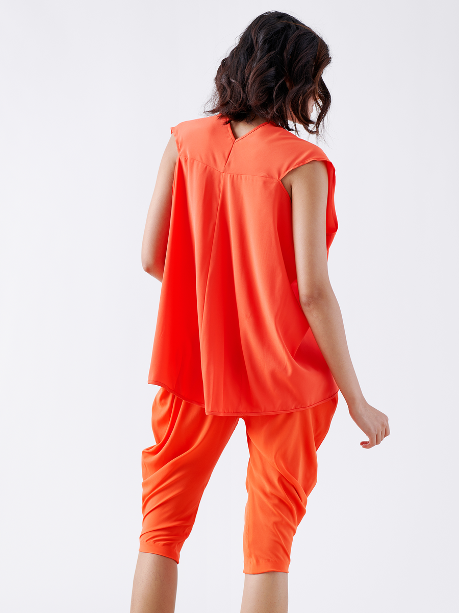 A person with wavy dark hair is wearing a bright orange sleeveless top and the Crop Curve Ori Pant from Zero + Maria Cornejo, featuring an elastic banded waist. They are standing with their back to the camera against a plain white background.