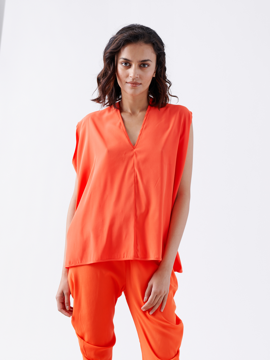 A woman with wavy, shoulder-length hair wears the Zero + Maria Cornejo Ina Tank and matching orange pants. She stands against a plain white background, looking directly at the camera with a neutral expression.