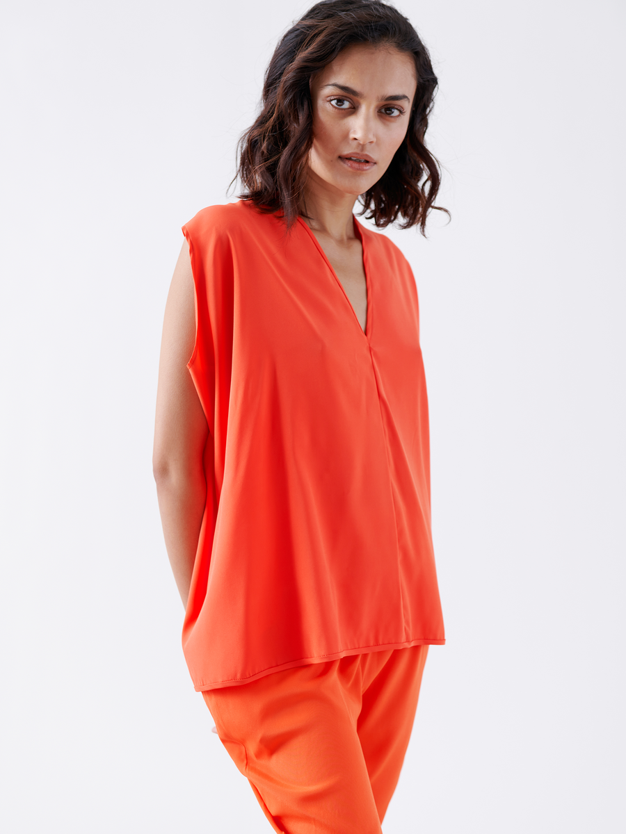 A person stands against a white background, wearing the vibrant orange Ina Tank from Zero + Maria Cornejo with matching pants. Their short, wavy hair frames a neutral expression, beautifully complementing the sleek silhouette of their attire.