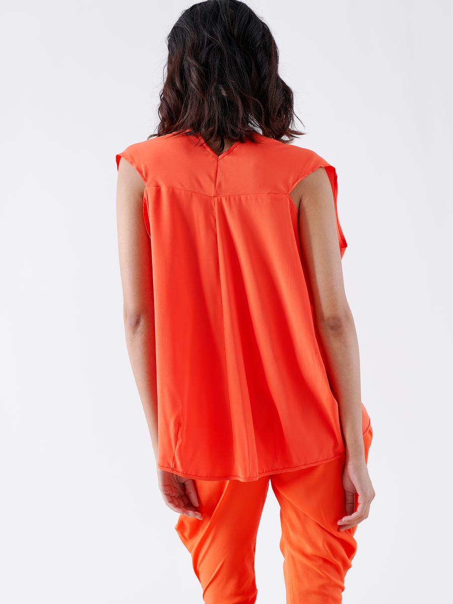 A person with shoulder-length dark hair wears the Zero + Maria Cornejo Ina Tank - Final Sale, a sleeveless, loose-fitting silk charmeuse top in vibrant orange with matching pants, creating a draped silhouette against a white background.