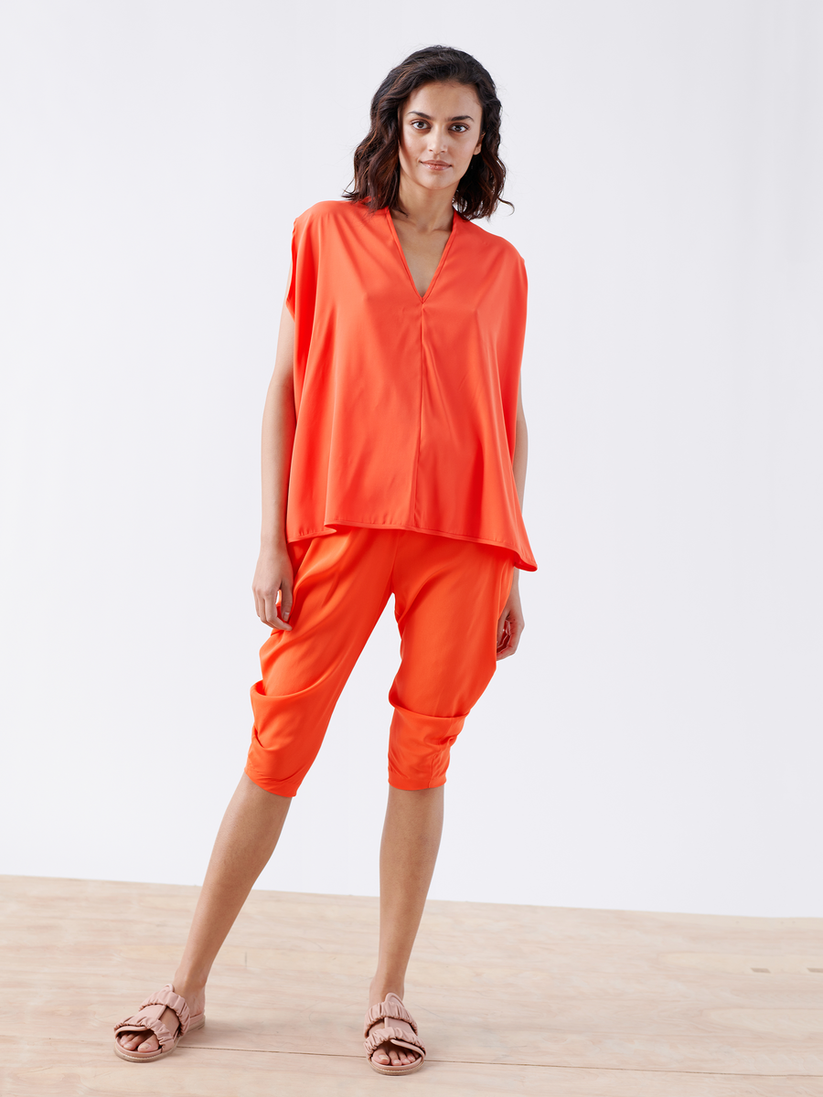 A woman in a vibrant orange ensemble, featuring Zero + Maria Cornejo's Ina Tank - Final Sale, stands on a wooden floor. Her pink sandals enhance her style as she poses against a plain white backdrop, exuding elegance with her draped silhouette.