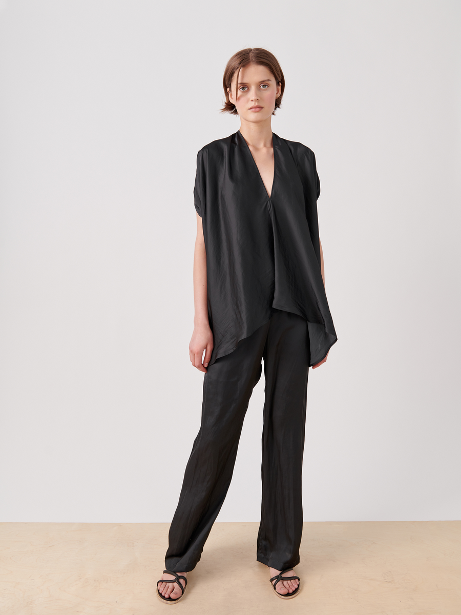 A person models the Gigi Pant from Zero + Maria Cornejo, wearing a flowing black top with short sleeves and matching wide-leg pants made of washable georgette. Against a plain white background, the ensemble is completed with black sandals.