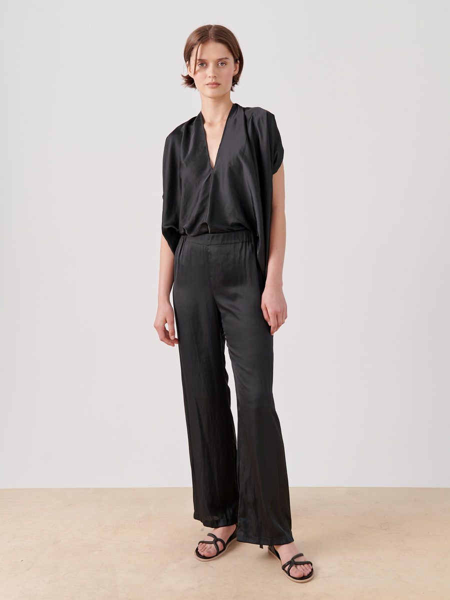 Against a plain backdrop, someone wears a flowy black outfit made from washable georgette. Featuring the Gigi Pant by Zero + Maria Cornejo, the ensemble includes short sleeves and wide-leg pants with a gathered waist, complemented by short hair and stylish black sandals.