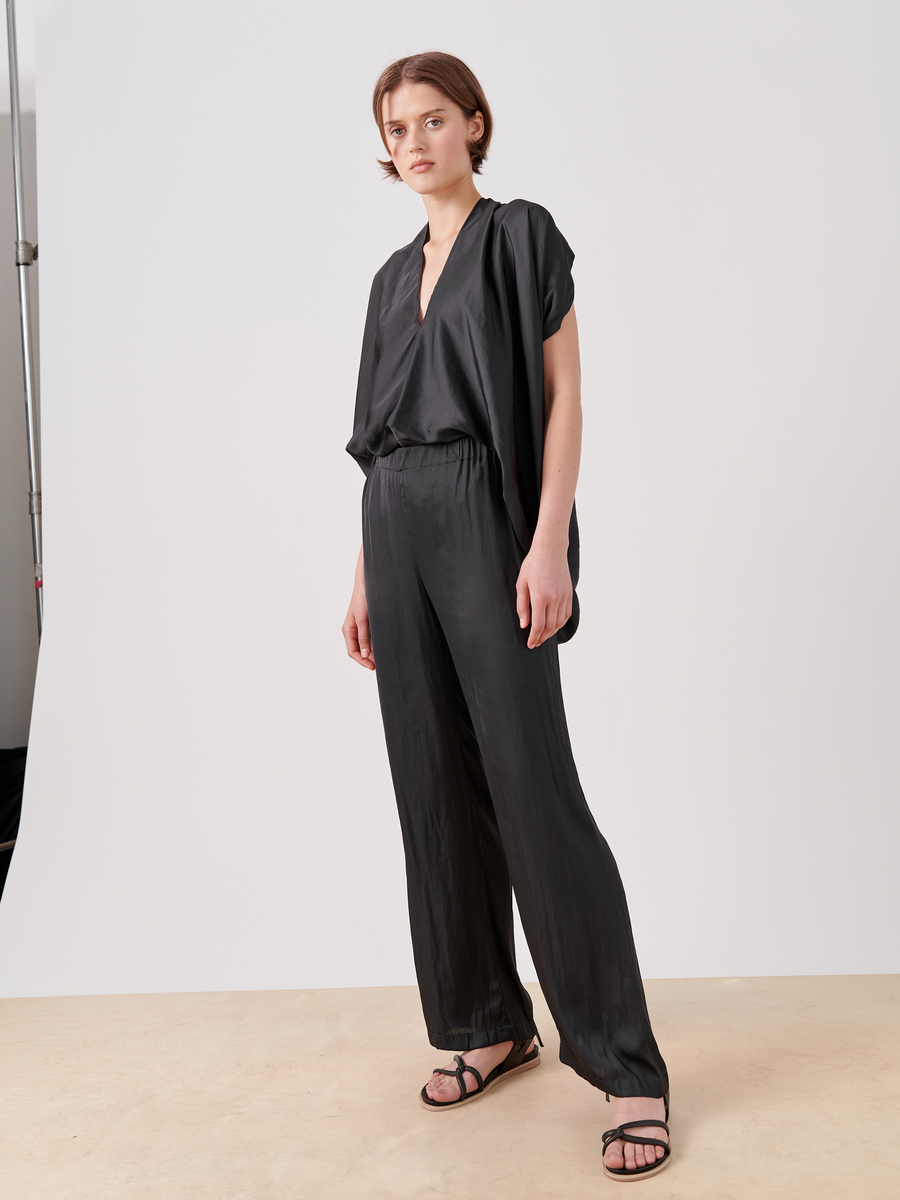 A person confidently stands against a plain white background, wearing a loose-fitting, flowing black outfit from Zero + Maria Cornejo. The ensemble features the Gigi Pant in washable georgette with wide legs. They have short brown hair and wear black sandals.