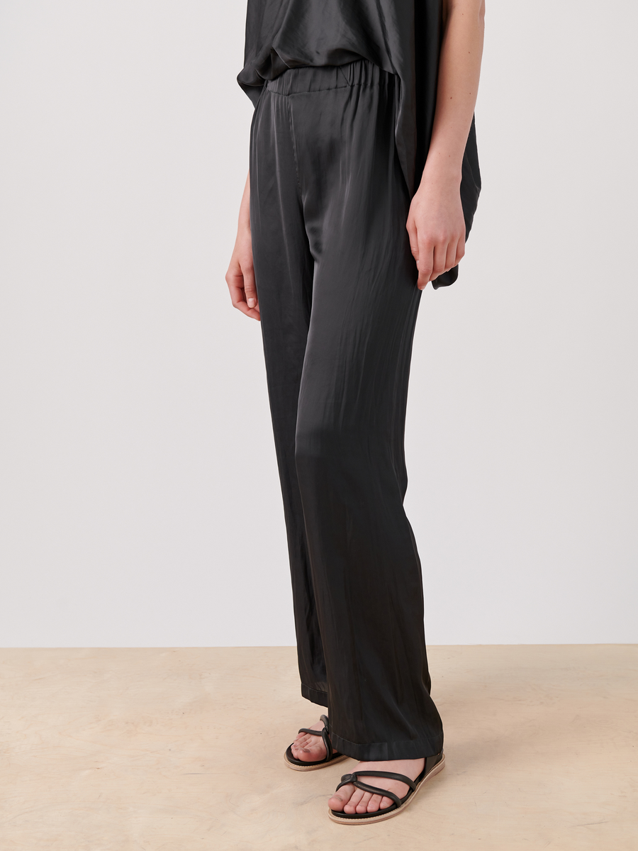 The person is wearing loose-fitting Gigi Pant - Final Sale by Zero + Maria Cornejo, featuring a gathered banded waist and a matching top. Black sandals complete the outfit. The floor is light-colored wood, with a clean, plain white backdrop.