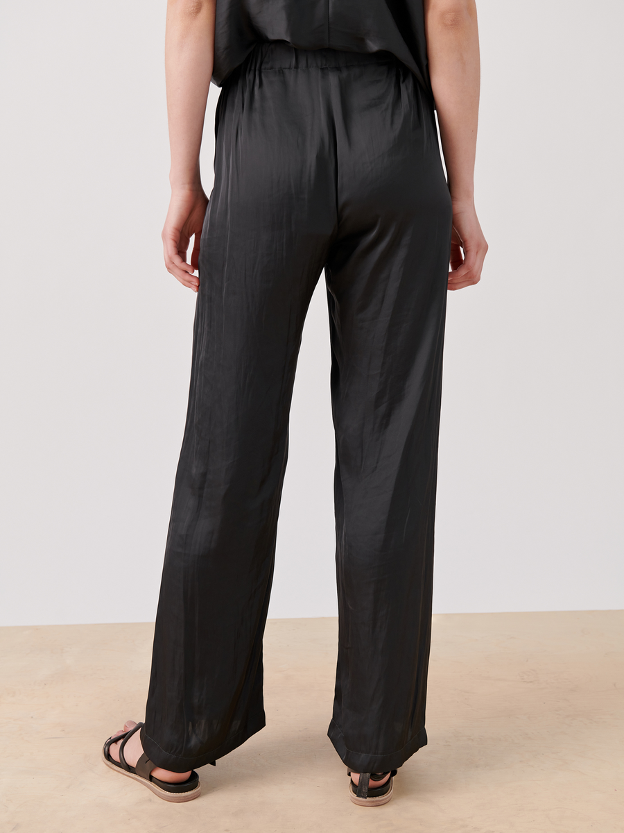 The person is wearing the Gigi Pant from Zero + Maria Cornejo, shown in black satin with an elastic waistband. They're standing on a light beige surface and wearing black sandals.