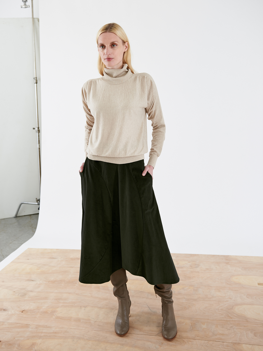 Someone wearing a beige turtleneck sweater and the Zero + Maria Cornejo Circle Swing Skirt - Final Sale in dark green stands on a light wooden floor against a plain white background, paired with knee-high brown boots.