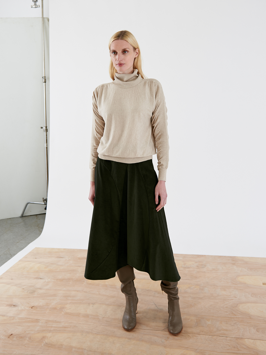 A person with long blonde hair stands on a wooden floor, dressed in a beige cotton elastane turtleneck sweater and the Zero + Maria Cornejo Circle Swing Skirt in dark green. They wear brown ankle boots, set against a plain white wall.