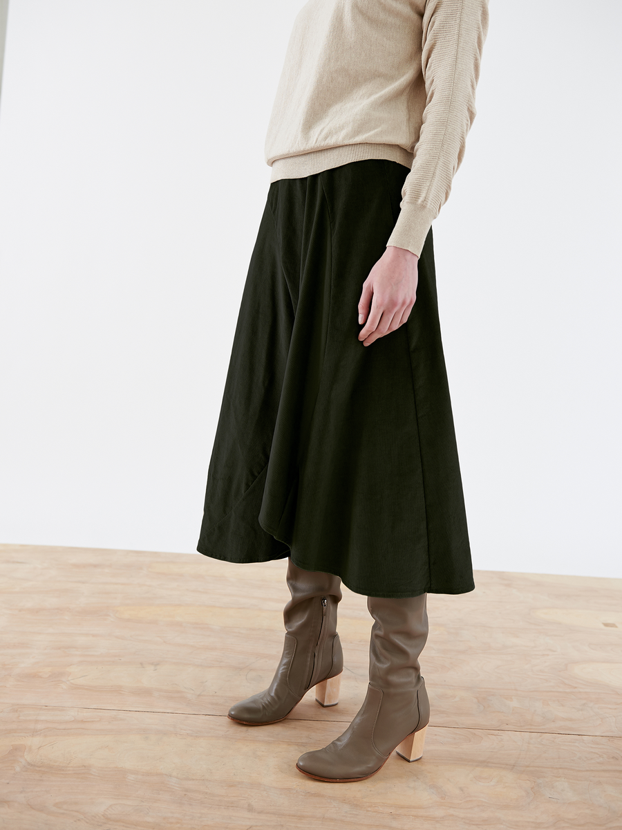 A person stands on a wooden floor against a white background wearing the Circle Swing Skirt - Final Sale by Zero + Maria Cornejo, an asymmetrical A-line dark green skirt, with a beige sweater and brown heeled boots. Their upper body is cropped out of the image.