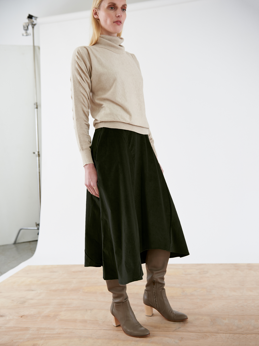 A person with long blonde hair wears a beige turtleneck sweater and the dark green Circle Swing Skirt by Zero + Maria Cornejo. They have gray heeled boots on, standing on a wooden floor against a plain white backdrop.