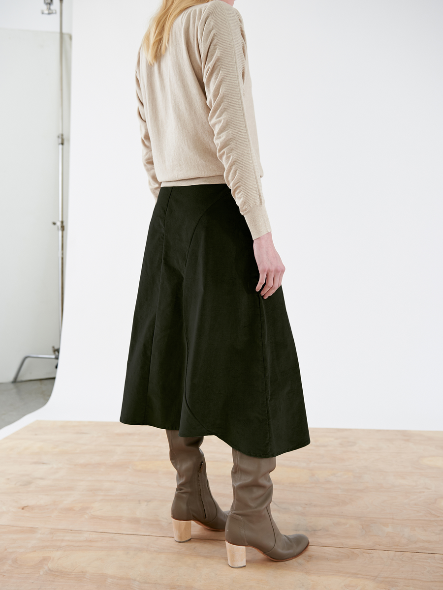 A person with long blonde hair is facing away, wearing a beige sweater, the Circle Swing Skirt - Final Sale from Zero + Maria Cornejo in dark green, and knee-high brown boots with block heels. They are standing on a wooden floor against a white backdrop.