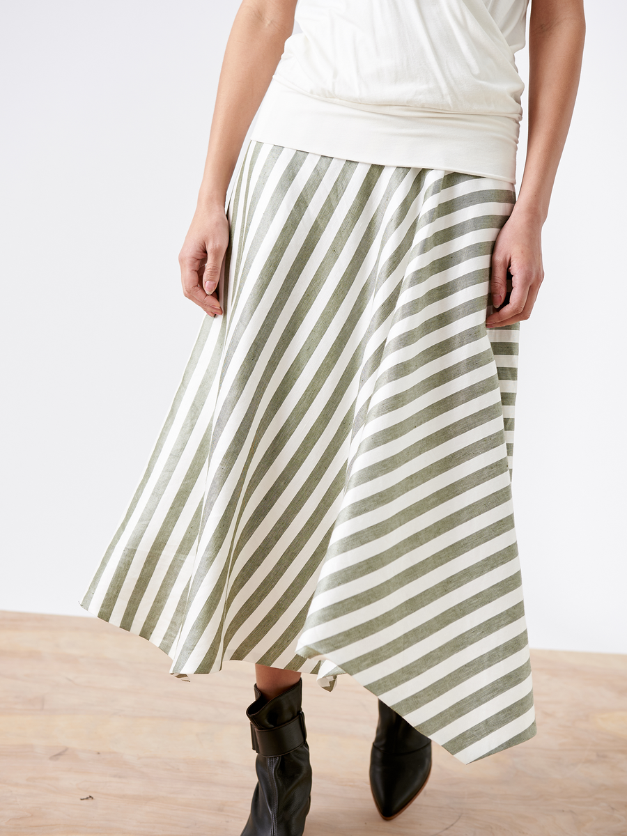 A person in a Zero + Maria Cornejo Aki Wave Skirt, featuring sustainable fabric with diagonal green and white stripes, pairs it with black ankle boots and a white shirt. They stand on a light wooden floor against a plain wall.