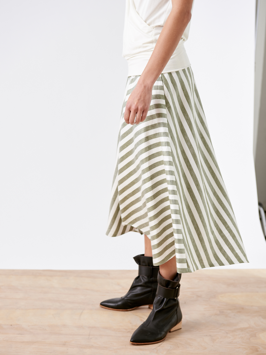 A person wearing a white top and the Aki Wave Skirt by Zero + Maria Cornejo—an asymmetrical midi skirt with green and white stripes, crafted from sustainable fabric—is paired with black ankle boots, standing on a wooden floor against a plain background.