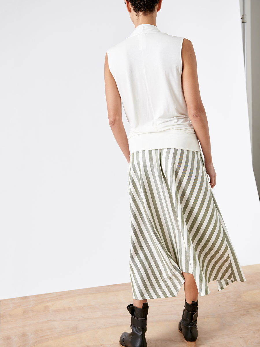 Facing away, a person wears a sleeveless white top with the Aki Wave Skirt from Zero + Maria Cornejo, featuring green and white stripes. They complete the look with black ankle boots. The plain white backdrop and wooden floor highlight the minimalist aesthetic.