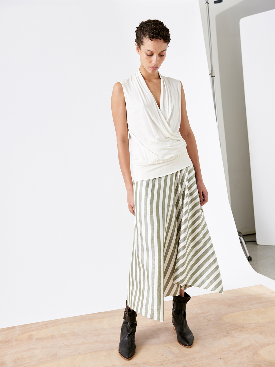 A person with short curly hair wears a sleeveless white wrap top and high-waisted green and white striped wide-leg Tencel fiber pants, paired with black ankle boots. They stand on a wooden floor against a plain white backdrop, not featured in the Aki Wave Skirt by Zero + Maria Cornejo collection.