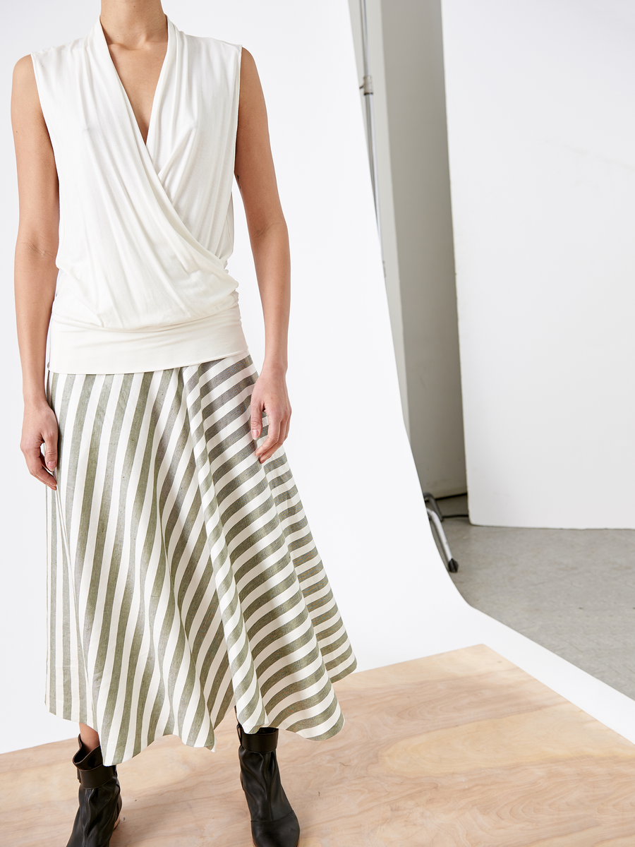 A person in a sleeveless white wrap top and the Aki Wave Skirt by Zero + Maria Cornejo, designed with sustainable fabric, stands on a wooden floor. Black ankle boots complete the outfit against a simple studio backdrop of white and gray elements.