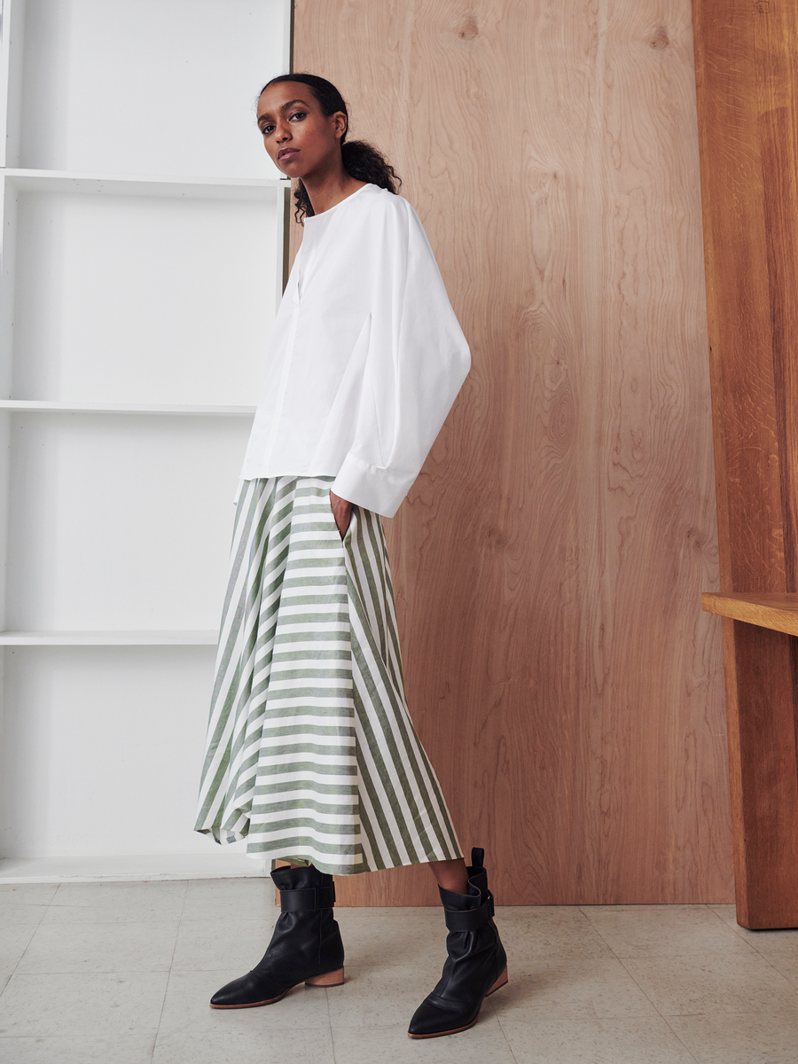 Inside a minimalistic setting with light wooden panels and white shelves, someone showcases a white blouse paired with the Aki Wave Skirt by Zero + Maria Cornejo. The sustainable Tencel fiber skirt features long stripes, complemented by black boots.