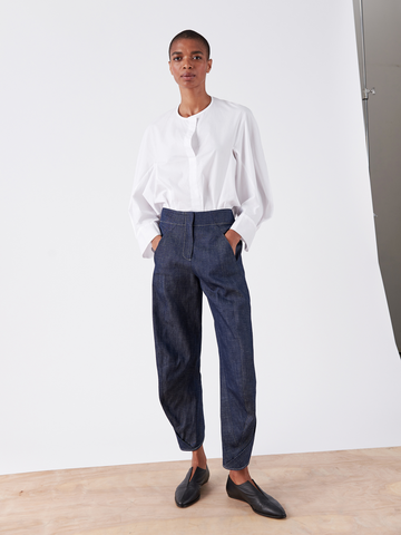 A person stands against a plain white background, dressed in a white long-sleeved, button-up shirt made of GOTS-certified cotton and the Takeo Pant by Zero + Maria Cornejo. They are wearing black slip-on shoes and have a neutral expression, with hands casually placed in their pockets.