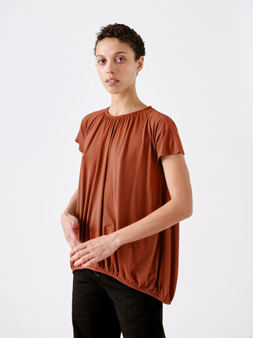 A person with short curly hair wears the rust-colored Girly Bubble Top by Zero + Maria Cornejo, paired with black pants. They stand in front of a plain white background, gazing at the camera with a neutral expression.