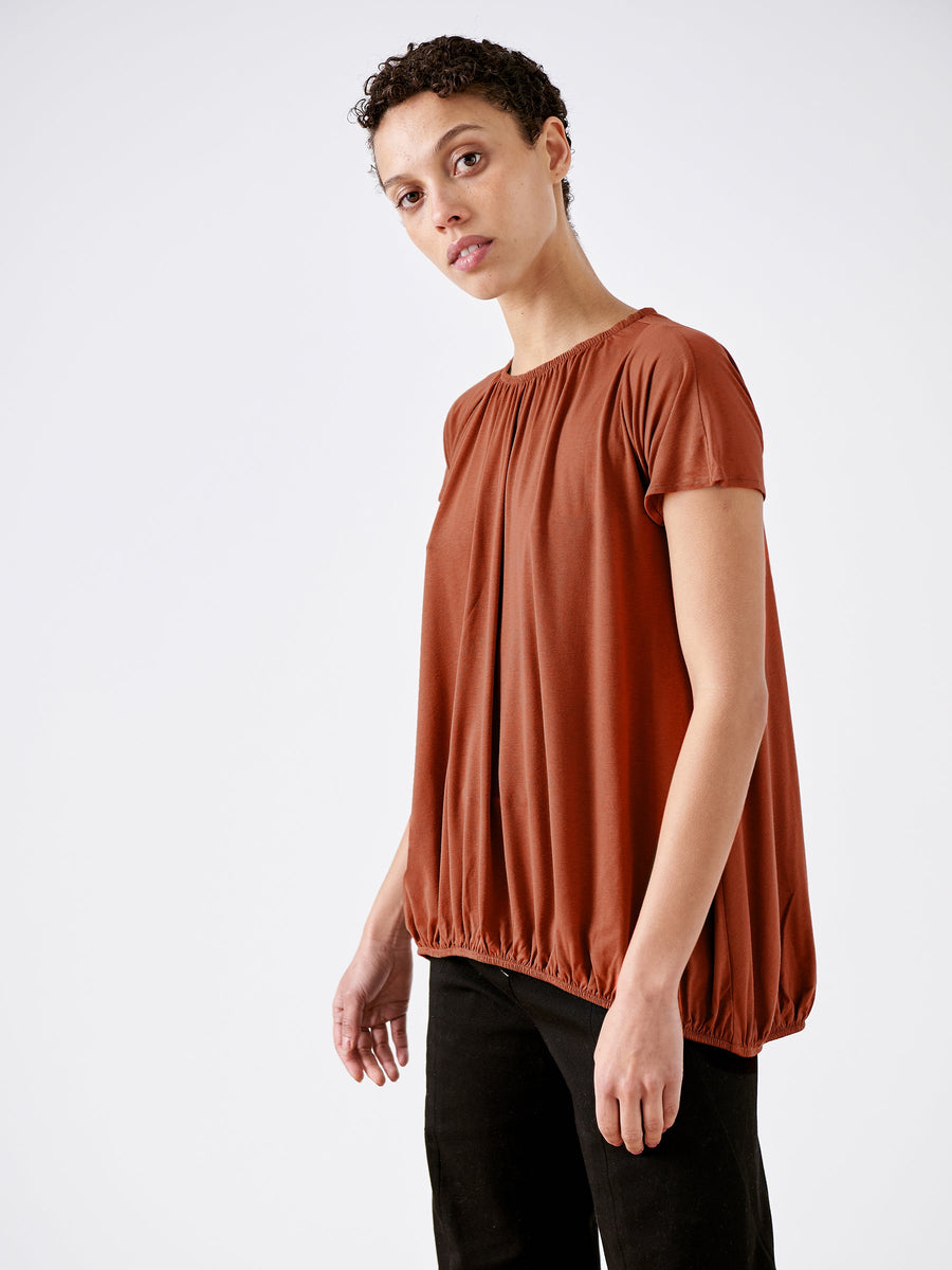 A person in a loose rust-colored Girly Bubble Top by Zero + Maria Cornejo and black pants stands against a plain white background, looking at the camera with a relaxed expression.