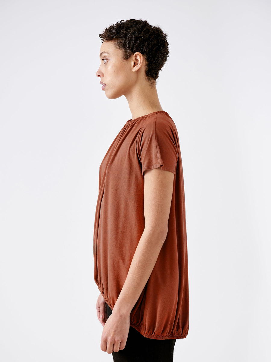 A person with short curly hair wears the Girly Bubble Top from Zero + Maria Cornejo, a loose-fitting, short-sleeved brown jersey crafted from soft viscose fibers, while standing in profile against a plain white background.