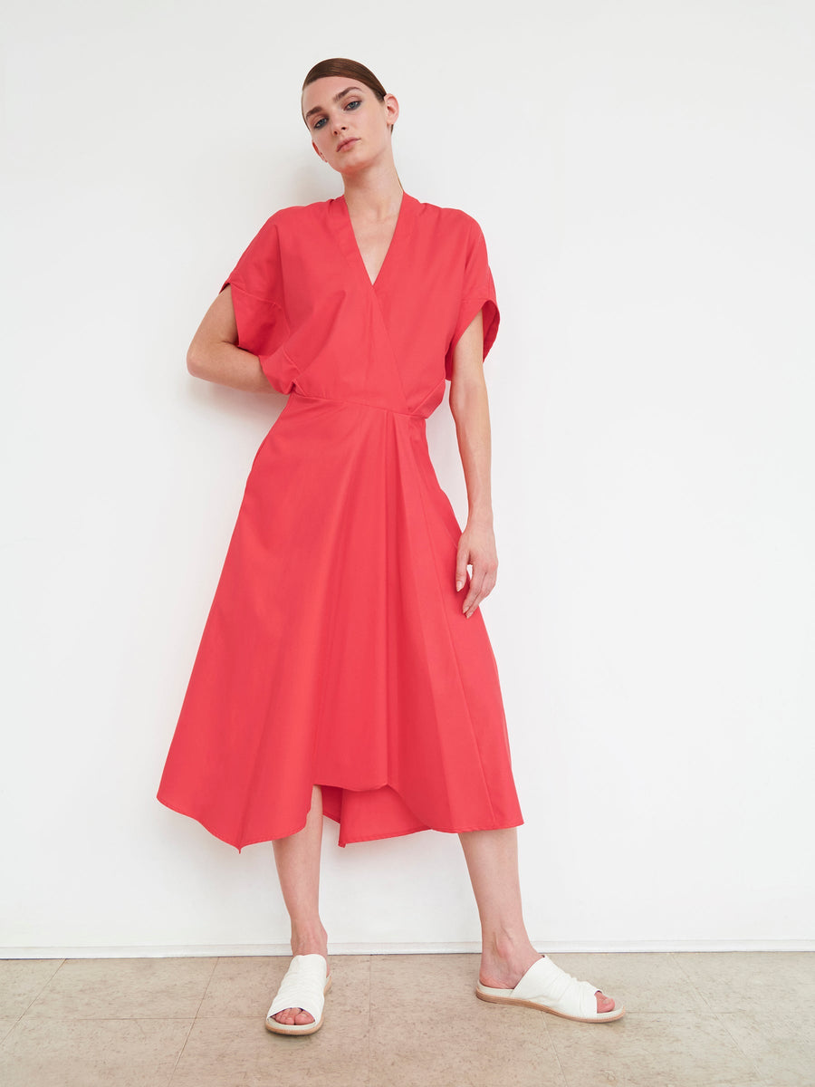 Standing against a white wall, a person wears the Aki Wave Dress by Zero + Maria Cornejo. This bright red cotton broadcloth dress has short sleeves, a V-neckline, an asymmetrical knee-length skirt, and is paired with flat white sandals. The person, with short hair, looks at the camera.