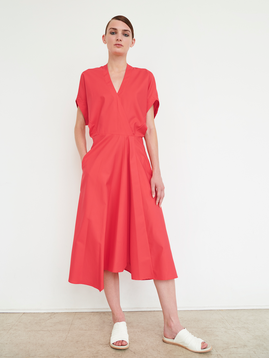 A person confidently poses against a white background wearing the Aki Wave Dress - Final Sale from Zero + Maria Cornejo. The bright red, knee-length wrap-effect dress features a V-neckline and pockets. They have short hair and wear white slide sandals.