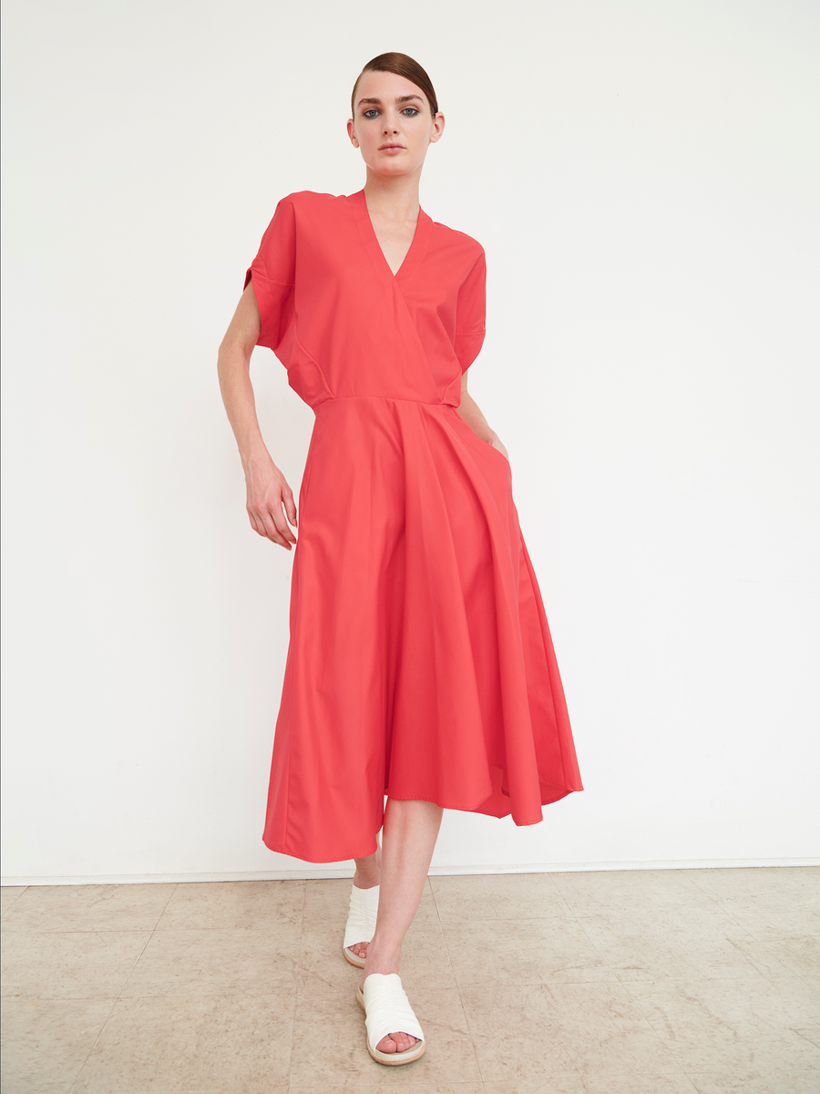 A person with short hair poses against a white background wearing the coral red Aki Wave Dress by Zero + Maria Cornejo, featuring a V-neck, knee-length skirt, short sleeves, pockets, and crafted from cotton broadcloth. The look is completed with white open-toe footwear.