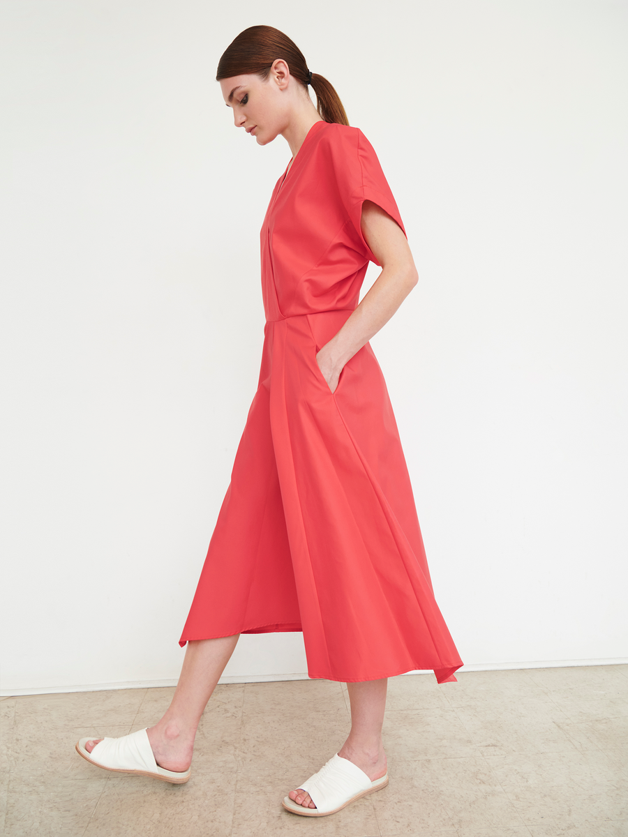 Dressed in the coral Aki Wave Dress by Zero + Maria Cornejo, featuring an asymmetrical skirt and paired with white sandals, a woman walks sideways with her hands in her pockets against a plain white backdrop, showcasing a minimalist and modern style.