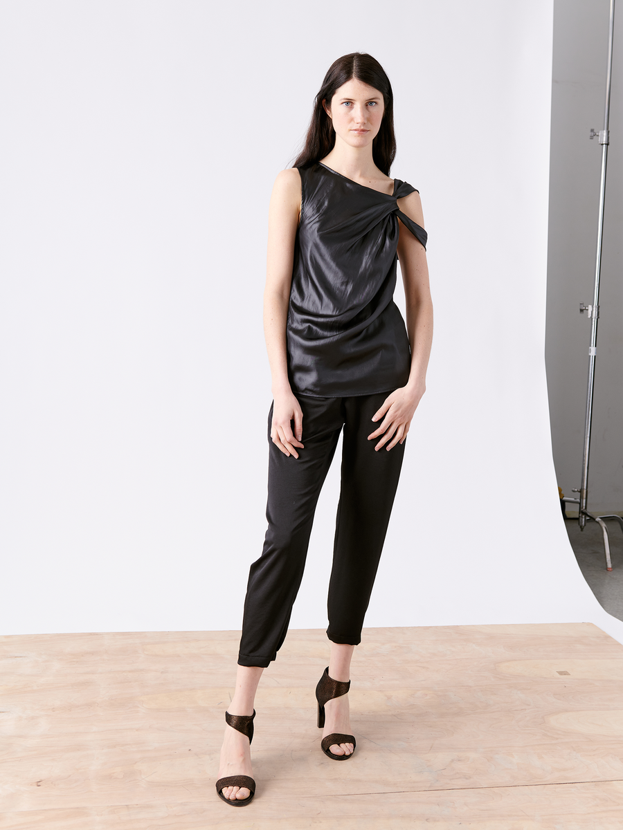 A woman with long dark hair stands on a wooden floor against a plain backdrop. She wears the stylish black Ampe Top by Zero + Maria Cornejo, made from recycled polyester, paired with black cropped pants and high heels. Her left hand is on her hip, and she maintains a neutral expression.