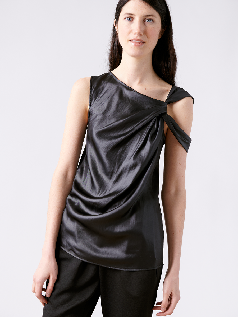 A person with long, dark hair wears the Ampe Top - Final Sale by Zero + Maria Cornejo, a shiny asymmetrical sleeveless top made from recycled polyester featuring a unique twisted design, paired with black pants. They smile slightly at the camera against a plain white background.