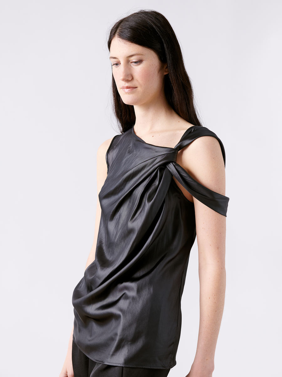 A person with long dark hair wears the stylish Ampe Top by Zero + Maria Cornejo. This black satin top features an asymmetric knot detail with one shoulder strap and elegant draping, set against a neutral light gray background.