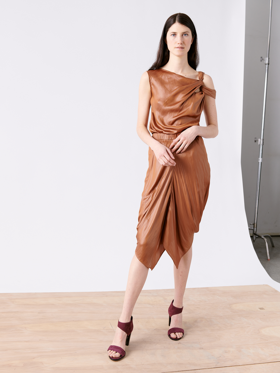 A woman stands on a wooden floor before a white backdrop, donning the Ampe Top by Zero + Maria Cornejo, featuring an elegant knot detail and asymmetrical sleeveless design in brown. She's paired it with maroon open-toe heels, her dark hair loose as she gazes at the camera.