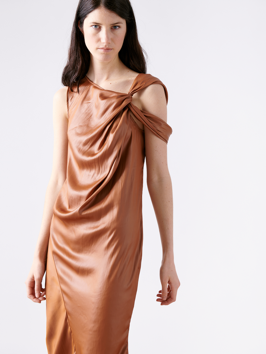 A person with long dark hair is wearing the Ampe Ibit Dress, a copper-colored cocktail dress by Zero + Maria Cornejo. Made from recycled polyester, it features draped material and an asymmetric shoulder knot. They stand against a plain, light background.