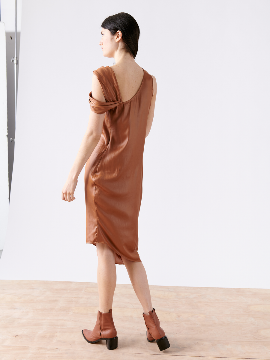 A person with dark hair stands on a wooden floor, wearing the Ampe Ibit Dress from Zero + Maria Cornejo and brown ankle boots. Facing sideways against a plain white background, the silky, sleeveless cocktail dress is made from recycled polyester.