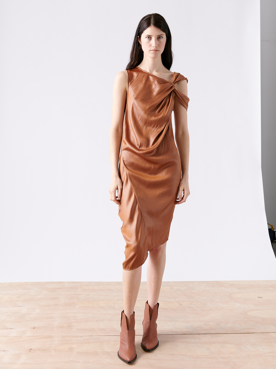 A woman with long dark hair stands on a wooden floor against a plain light wall, wearing Zero + Maria Cornejo's Ampe Ibit Dress. This brown cocktail dress boasts an asymmetrical, one-shoulder design with an elegant knot and is made from recycled polyester, perfectly paired with brown ankle boots.