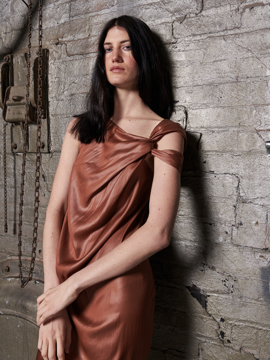 A person with long dark hair stands confidently against a textured brick wall, wearing the Ampe Ibit Dress from Zero + Maria Cornejo. Made of recycled polyester, this brown cocktail dress features an asymmetric knot, with chains adorning the wall complementing their crossed arms and allure.