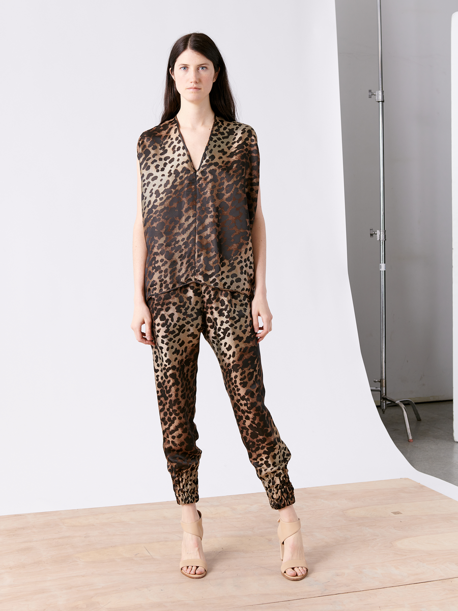 A person with long dark hair wears the sustainably-sourced Ina Tank and leopard print pants by Zero + Maria Cornejo, standing on a wooden floor against a plain studio backdrop, accessorized with nude high-heeled sandals.