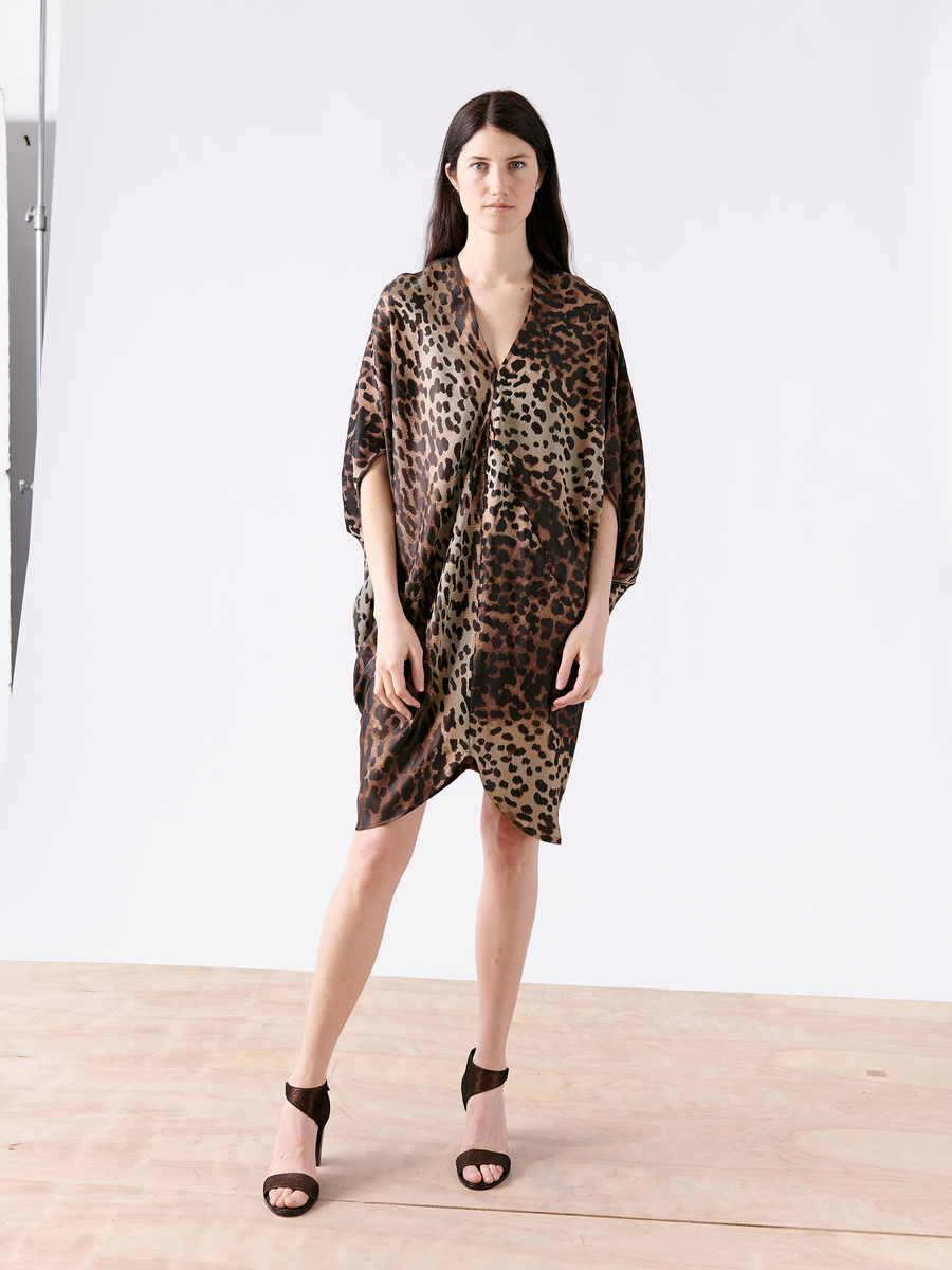 An individual stands on a wooden floor wearing a loose, cocoon-shaped Koya Dress by Zero + Maria Cornejo, featuring a leopard-print design in sustainably-sourced viscose, paired with black high-heeled sandals. The background is a plain white wall.