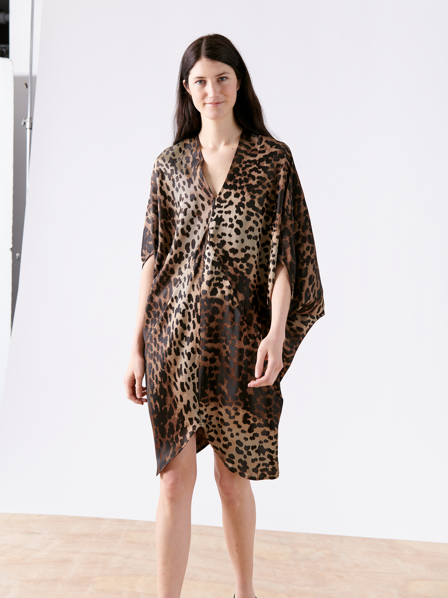 A person with long dark hair wears the Koya Dress by Zero + Maria Cornejo, showcasing a loose-fitting cocoon silhouette and leopard print in sustainably-sourced viscose. They smile slightly, standing on a light wooden floor against a white background.