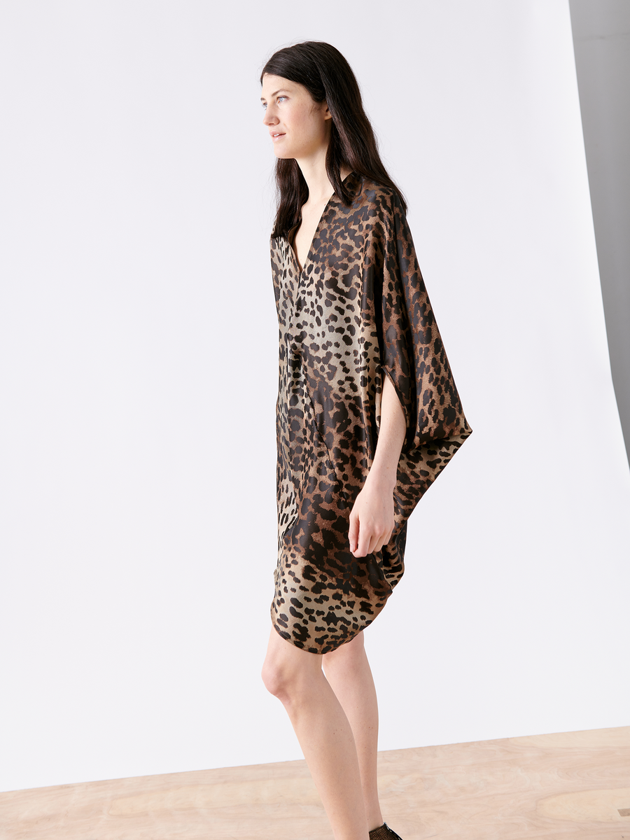 A person with long dark hair wears the Koya Dress from Zero + Maria Cornejo, standing on a wooden floor against a white backdrop. The leopard-print cocoon silhouette in sustainably-sourced viscose drapes elegantly as they look left, their right arm relaxed by their side.