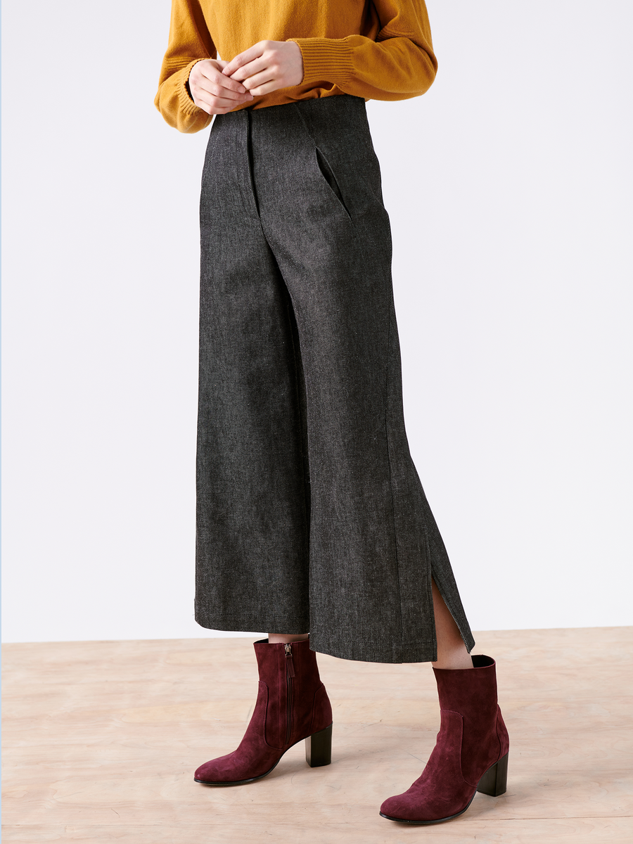 A person wearing a Cropped Fin Pant from Zero + Maria Cornejo, in gray traceable denim, paired with a mustard sweater and burgundy block-heeled ankle boots, stands on a light wood floor against a plain white background.